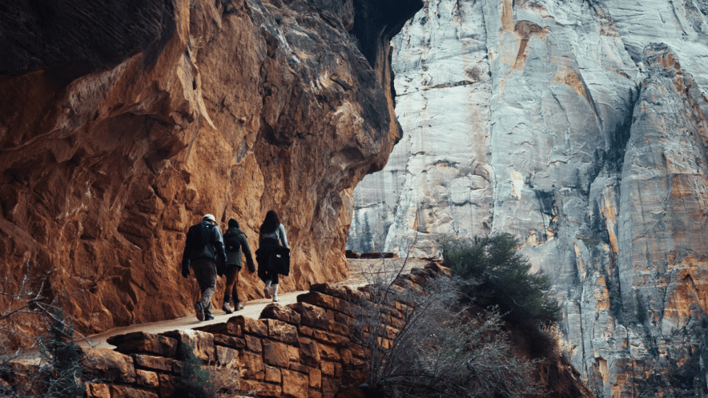Three hikers walk along a narrow, rocky path carved into the side of a towering cliff at Scout Lookout in Zion National Park, with sheer rock faces rising dramatically in the background.