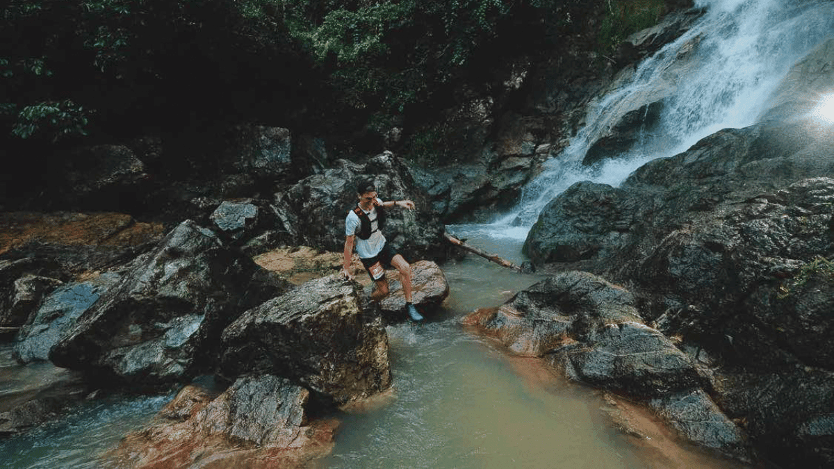This image shows a person running through a rocky, lush forest area near a waterfall. The hiker is stepping on a large rock surrounded by flowing water, with the waterfall cascading down rocks in the background, and dense green foliage framing the scene. The image captures an adventurous outdoor activity in a natural setting.