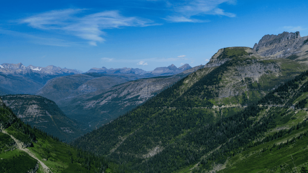 A panoramic view from the start of the Highline Trail in Glacier National Park. It shows vast mountainous terrain featuring layers of green valleys, forested ridges, and distant snow-capped peaks beneath a clear blue sky with light wisps of clouds. The lush greenery contrasts against the rocky mountain slopes, suggesting a high-altitude hiking area.