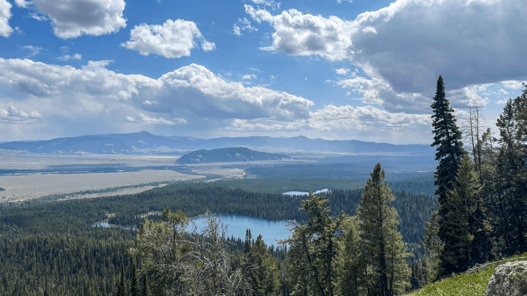 A scenic overlook showing Bradley Lake and Taggart Lake, surrounded by dense pine forests. In the distance, mountains rise under a partly cloudy sky with rolling clouds casting shadows on the valley below.
