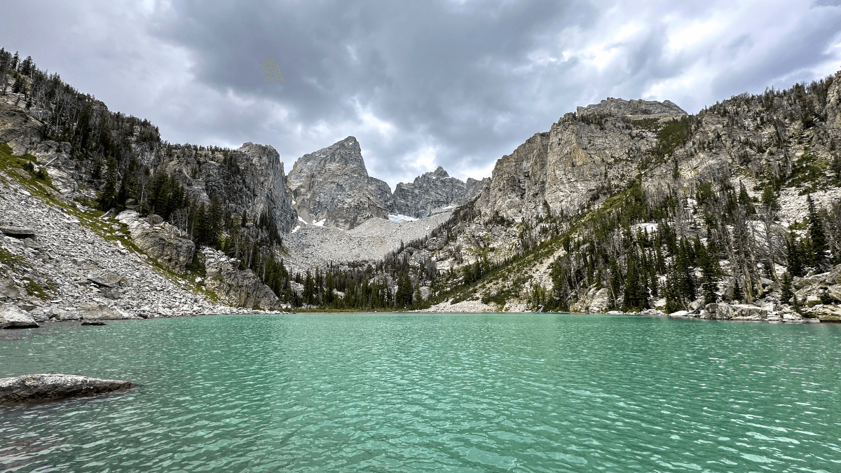 This image shows the alpine lake with bright turquoise water, surrounded by steep, rocky mountains and patches of evergreen trees. The sky above is overcast, with thick clouds casting a moody ambiance over the landscape. The image evokes a peaceful yet rugged wilderness setting. The lake comes at the end of the Delta Lake Trail in Grand Teton National Park.