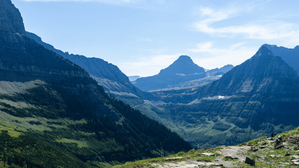 A sweeping view of jagged mountain peaks and deep valleys along the Highline Trail, leading toward Haystack Butte. The foreground shows a grassy, rocky trail with a couple of hikers visible in the lower right corner, while the backdrop features towering mountains and lush green ridges under a bright sky.