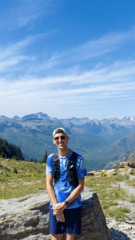 A man standing in front of a large rock, smiling while wearing sunglasses and a blue hiking outfit with a vest. Behind him, the expansive view of a mountainous valley unfolds with rugged peaks in the distance and a clear blue sky above.