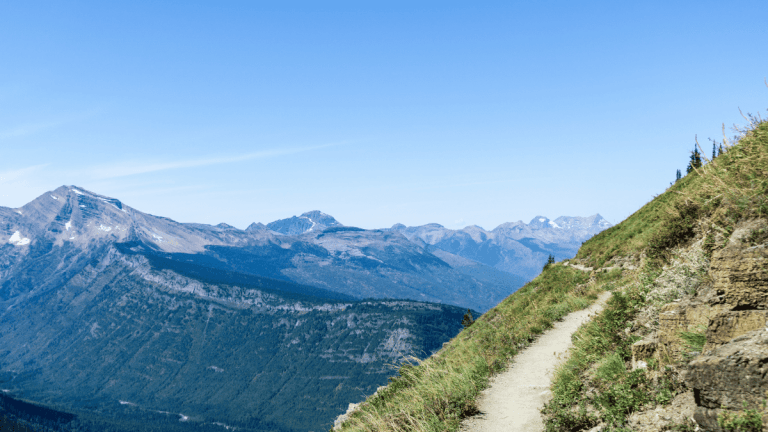 A narrow hiking trail winding along a steep mountainside, with expansive views of a rugged mountain range in the distance. The trail hugs the edge of the grassy slope, offering hikers a scenic path with a sweeping backdrop of jagged peaks and deep valleys.