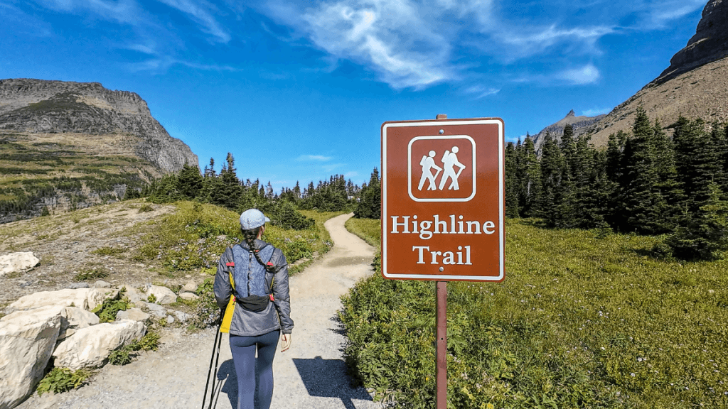 A hiker with trekking poles walks along a dirt path toward a trailhead, with a prominent brown sign marking the "Highline Trail." The surrounding landscape is filled with greenery, mountains, and a bright blue sky overhead.