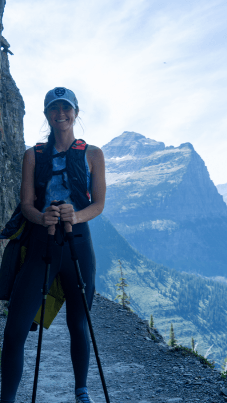 A woman standing on a narrow trail with hiking poles, wearing a hat, vest, and athletic gear. She is smiling, with towering mountain peaks visible in the background and soft clouds in the sky, showcasing the scenic beauty of the Highline Trail.