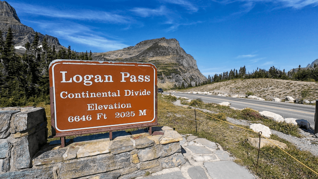 A large brown sign at Logan Pass, marking the Continental Divide at an elevation of 6,646 feet (2,025 meters). The sign is positioned in front of a stone base, with the road and towering mountain peaks visible in the background under a clear blue sky.