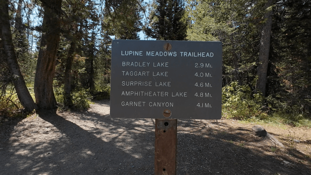 A metal sign at the Lupine Meadows Trailhead provides distances to various destinations: Bradley Lake (2.9 miles), Taggart Lake (4.0 miles), Surprise Lake (4.6 miles), Amphitheater Lake (4.8 miles), and Garnet Canyon (4.1 miles).
