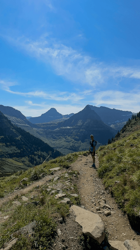 A trail runner pauses on a narrow, rocky path of the Highline Trail, overlooking a vast landscape. Behind him, majestic mountain peaks and verdant valleys stretch under a clear blue sky, dotted with fluffy clouds.