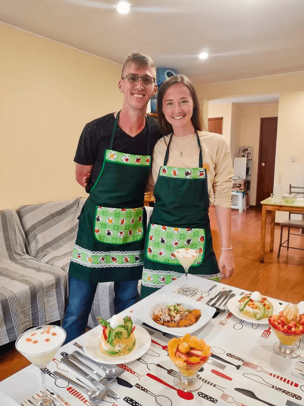 A smiling couple stands in a cozy dining room, both wearing matching green aprons with fruit designs. In front of them is a table set with an array of colorful dishes, including a martini glass dessert, a salad, and other plated foods. The background features a sofa and bright lighting.