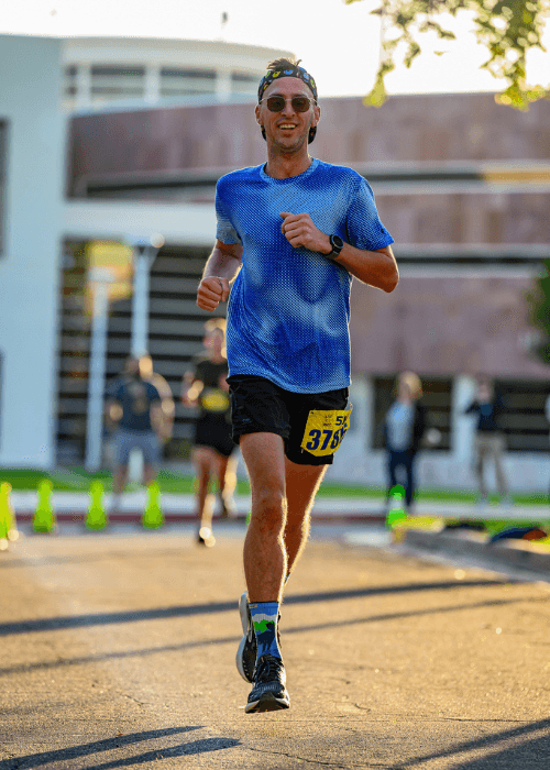 This image shows The Travel Runner mid-run in a bright, outdoor setting, likely during a race or an organized run. The runner is wearing sunglasses, a blue running shirt, and black shorts, with a race bib numbered "315." The background includes a modern building and other participants, slightly out of focus, with traffic cones marking the course. The runner appears to be smiling and in good spirits as they continue on their path.