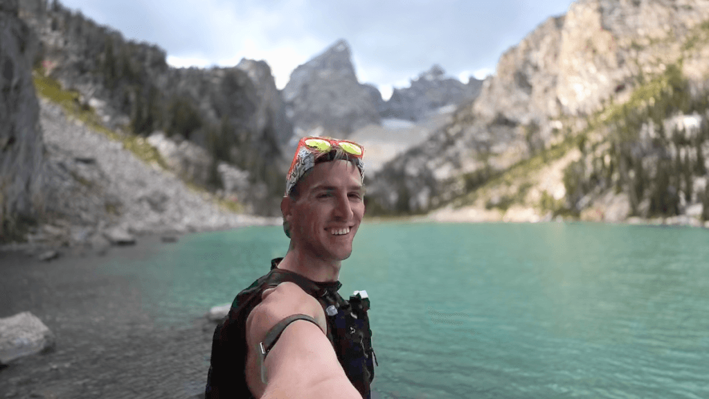 A man takes a selfie at a serene mountain lake with teal-blue water, surrounded by rugged, rocky cliffs and pine trees. Snow-capped peaks rise in the background under a partly cloudy sky, capturing a moment of adventure and natural beauty.