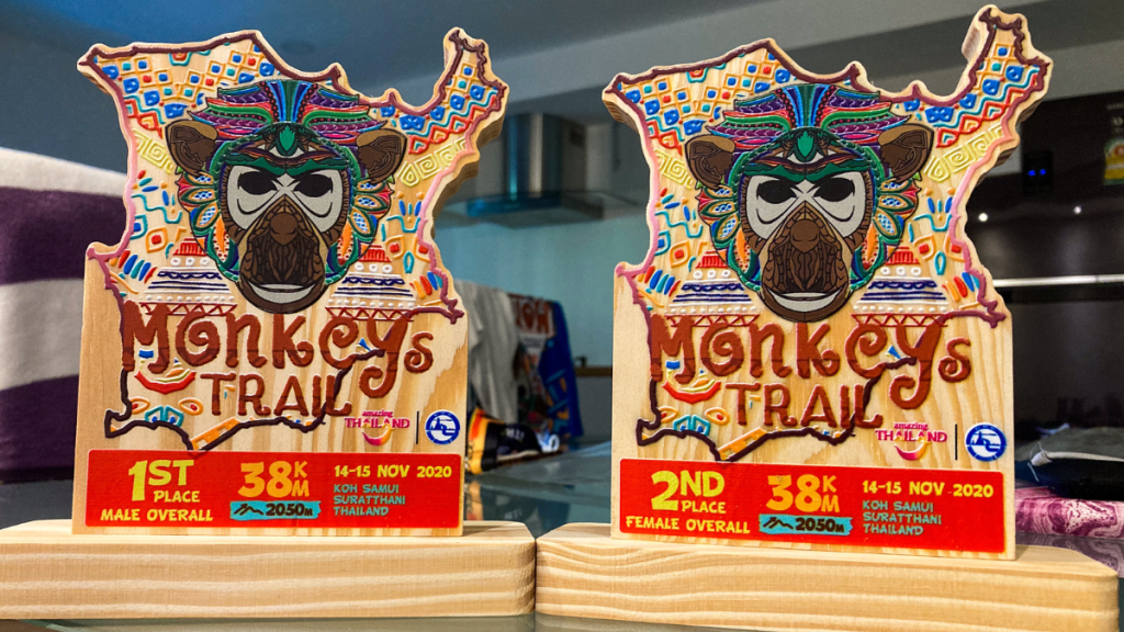 This image shows two colorful wooden trophies from the "Monkey's Trail" race, featuring a vibrant tribal-style monkey design and intricate patterns. The trophies commemorate a 38 km trail run held on November 14-15, 2020, in Koh Samui, Surat Thani, Thailand, with distinctions for "1st Place Male Overall" and "2nd Place Female Overall." The vibrant designs and event branding emphasize the unique and adventurous spirit of the race.