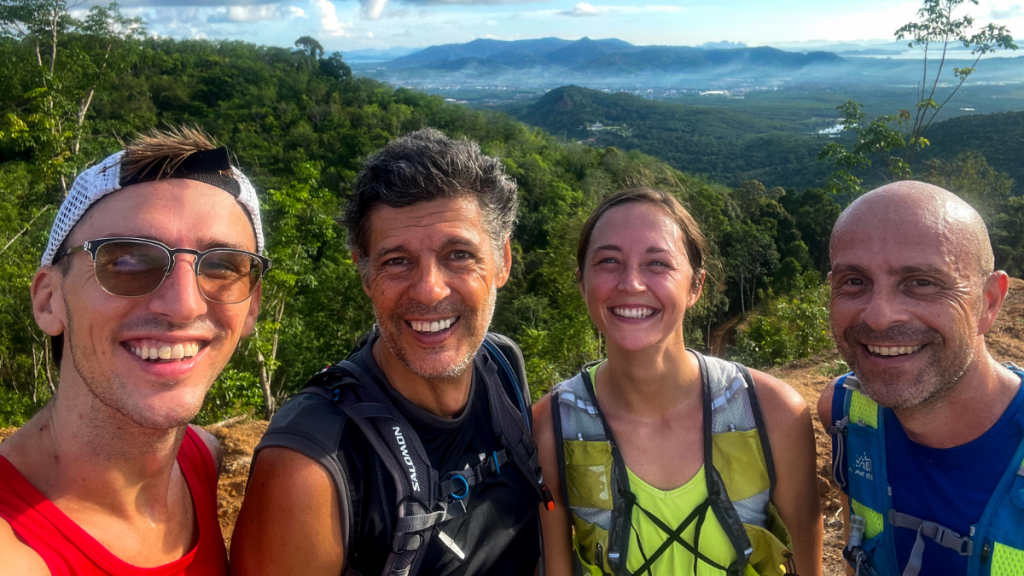 This image captures a group of four smiling trail runners posing for a selfie atop a lush green hill, with expansive views of valleys and mountains in the background. Each person is equipped with hydration packs, indicating they are mid-activity during a scenic run or hike. The vibrant greenery and clear sky emphasize the outdoor adventure and camaraderie shared among the group.