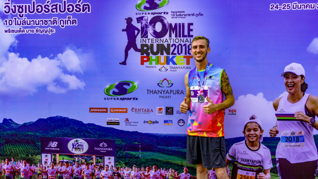 This image depicts a participant at the Supersports 10 Mile International Run in Phuket, 2018. The man stands proudly in front of the event backdrop, wearing a vibrant running jersey and displaying his race medal, with promotional logos and text in English and Thai. Smiling runners, including a child, can be seen in the background, conveying a festive and athletic atmosphere.
