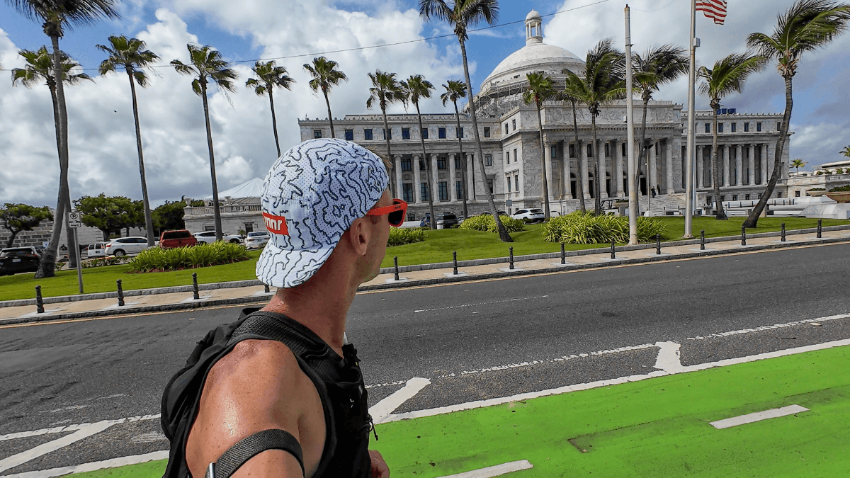 A runner stands on a street in front of the Puerto Rico Capitol building, wearing a white patterned cap and orange sunglasses, looking up at the historic structure. Tall palm trees and American flags line the path, adding a tropical vibe to the urban setting.