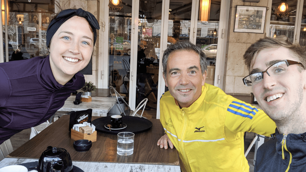 Three friends smile at the camera while seated at an outdoor cafe table, with coffee cups and a teapot in front of them. One wears a purple jacket and headband, another sports a yellow Mizuno jacket, and the third wears glasses, capturing a casual, friendly meetup. Local running tours are a great way to fit in running and sightseeing on a trip.