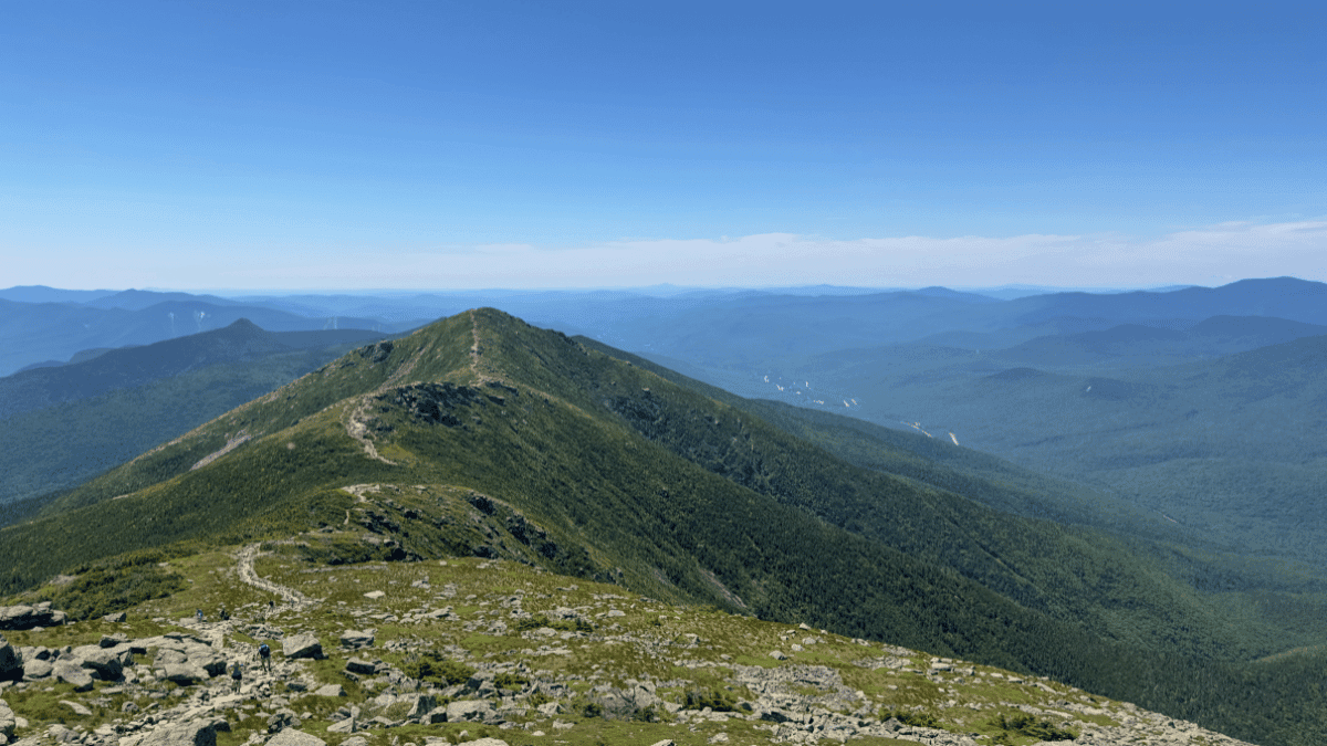 A scenic view of a rugged hiking trail winding along a green mountain ridge in White Mountain National Forest under a clear blue sky. The trail stretches toward distant peaks, surrounded by layers of forested mountains fading into the horizon.