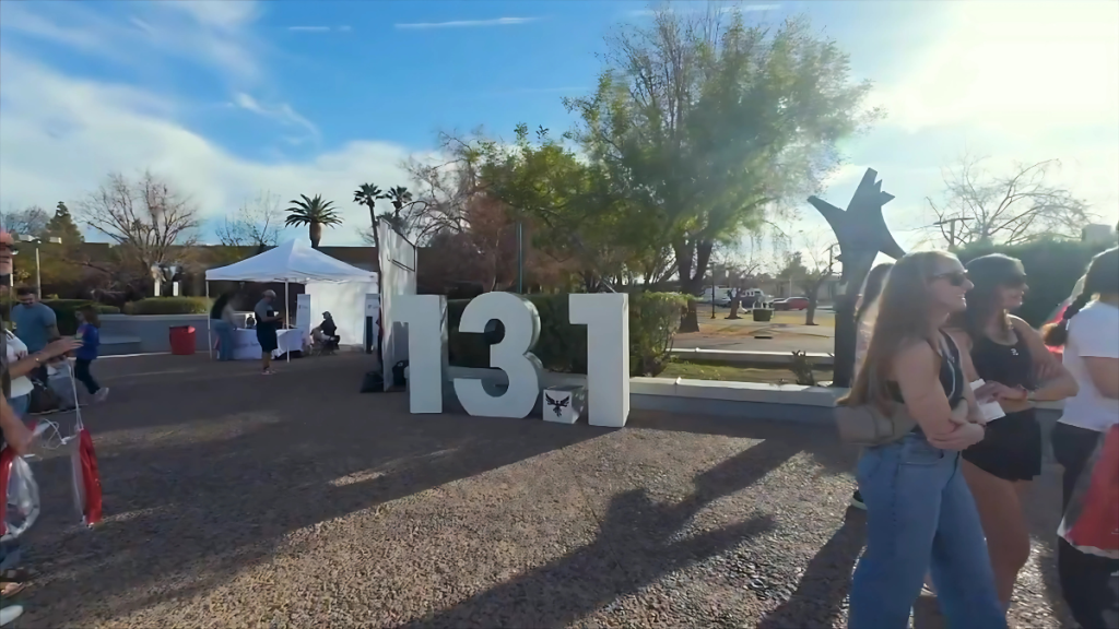 A large outdoor Mesa Half Marathon event sign with bold white numbers "13.1" stands on a paved area at the Mesa Half Marathon. Several people are gathered nearby, with white tents and a blue sky in the background.