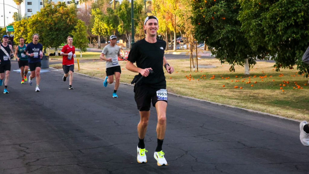 A smiling male runner in a black athletic outfit and white cap runs past orange trees on a park pathway during the Mesa Half Marathon. Other runners follow behind him, and fallen oranges are scattered on the grass.