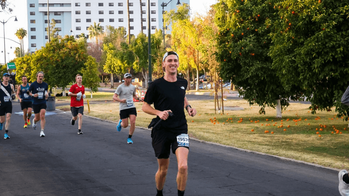 A male runner wearing a black athletic outfit and white cap runs on a scenic road during the Mesa Half Marathon. He smiles while holding a camera on a stabilizer. The background features lush green trees, a group of runners, and a clear blue sky.
