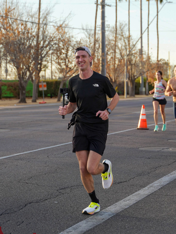 A male runner in a black North Face shirt, black shorts, and neon yellow running shoes runs on an open road during the Mesa Half Marathon. He smiles while holding a small camera on a stabilizer. Orange traffic cones and other runners are visible in the background.