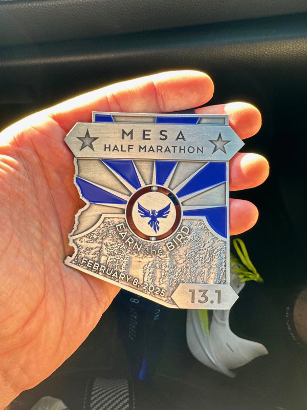 A close-up of a hand holding a Mesa Half Marathon finisher's medal. The medal is shaped like the state of Arizona, featuring blue accents, an engraved phoenix, and the words "Earn the Bird" with the date "February 8, 2025." A running shoe is slightly visible in the blurred background.