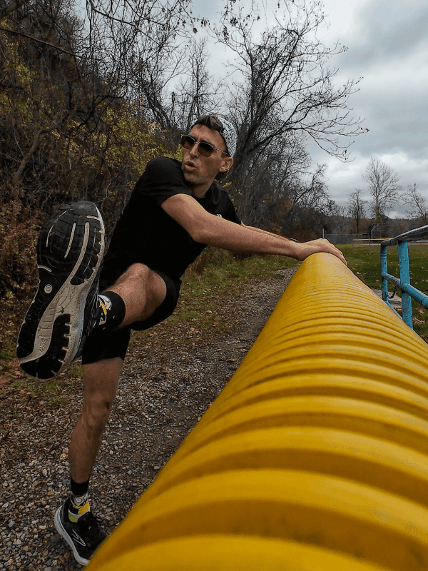 A man in athletic gear and sunglasses swings one leg over a bright yellow rail during a warm-up or stretching routine on a gravel trail. He’s mid-motion with a focused expression, surrounded by leafless trees under a cloudy sky.