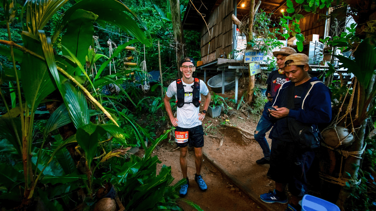 A smiling trail runner wearing a white shirt, hydration vest, and race bib 18990 stands on a dirt path surrounded by lush tropical plants, with two men nearby checking a phone. A wooden sign behind him points toward a waterfall 200 meters away and a coffee shop, hinting at a mid-run break in a jungle setting.
