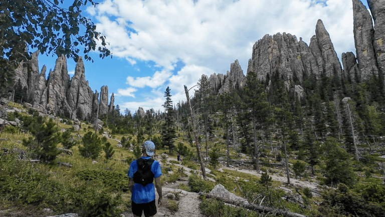 A hiker in a blue shirt and patterned cap walks along a dirt trail surrounded by rugged pine forest and towering, jagged rock spires. The sky is partly cloudy, casting a dramatic light over the striking geological formations, creating a sense of awe and adventure.