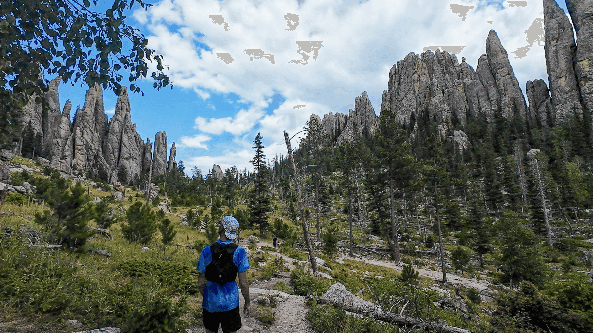 A hiker in a blue shirt and patterned cap walks along a dirt trail surrounded by rugged pine forest and towering, jagged rock spires. The sky is partly cloudy, casting a dramatic light over the striking geological formations, creating a sense of awe and adventure.