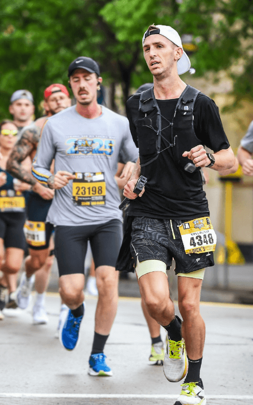 A group of runners in the marathon includes a man in the foreground, focused and striding forward, with bib 4348 and a white hat turned backward. Runners behind him appear in motion blur.