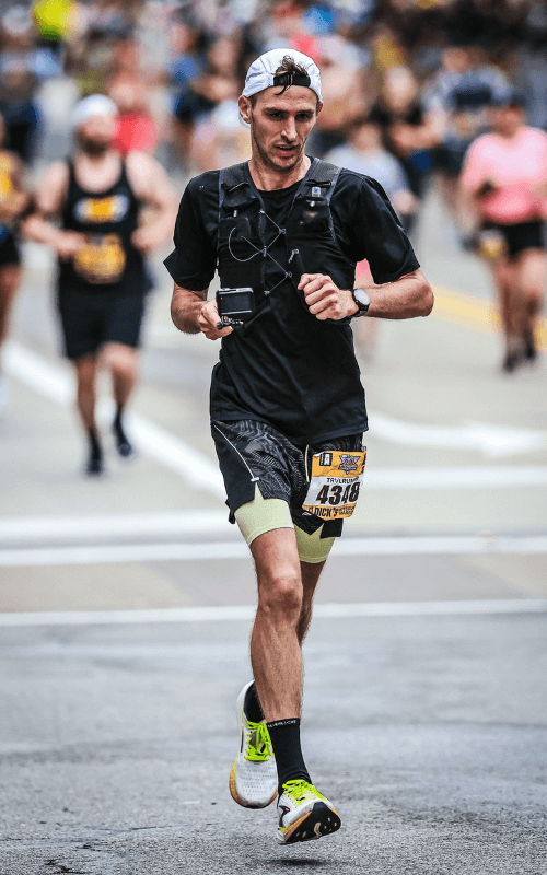 A male runner with a backwards white cap and black race gear is mid-stride during a marathon, with a determined look and other runners blurred in the background on a city street.