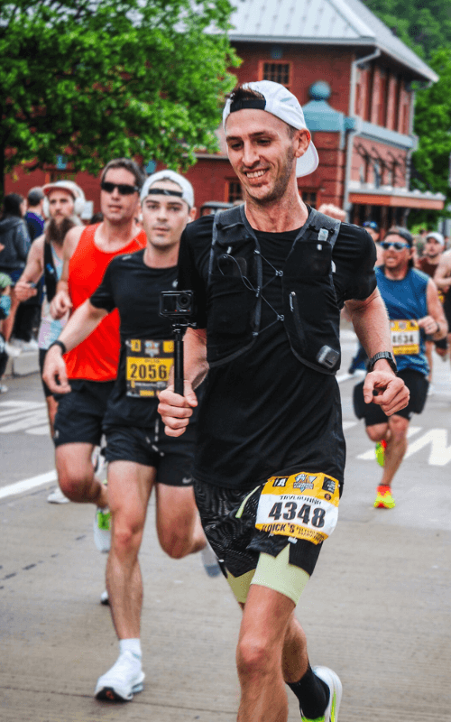 A smiling male runner holds a steady pace early in the race, surrounded by other participants. He’s wearing bib 4348 and a GoPro mounted on his chest.
