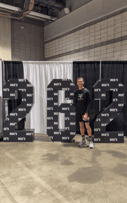 A man stands inside a convention center posing with a large “2025” display decorated with DICK’S logos, wearing running gear and smiling.