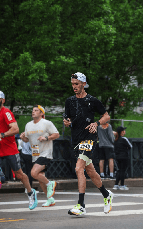 A runner in a backwards cap and hydration vest runs along a tree-lined street with several runners behind him. He looks down slightly, maintaining pace mid-marathon.