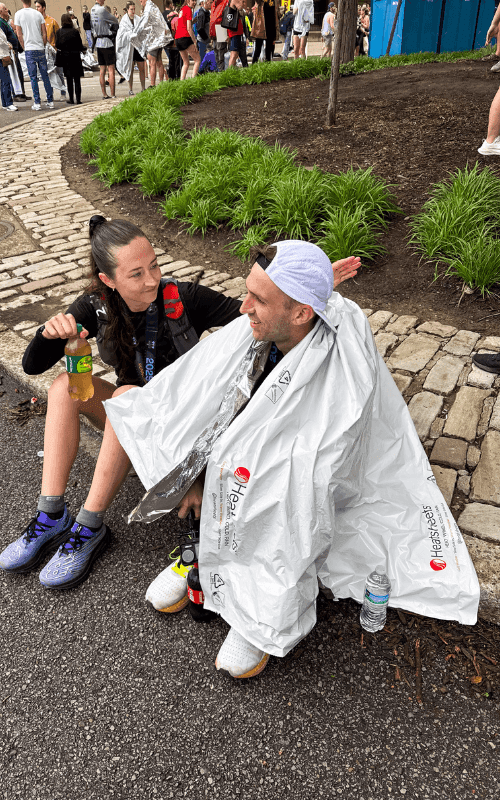 Two runners sit on the pavement near a landscaped area post-race, one covered in a silver finisher’s blanket and the other holding a green juice bottle, both smiling and sharing a moment of rest.