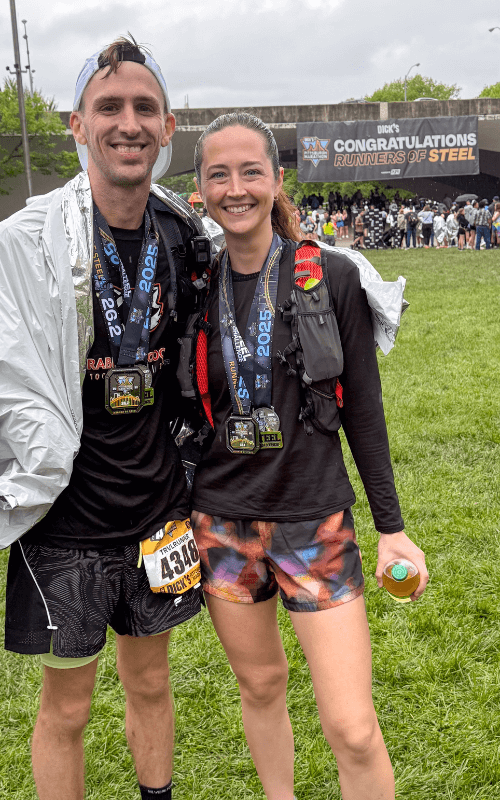 A man and woman stand smiling with arms around each other after a race, both wearing black running gear, finisher medals, and race bibs. Behind them is a large banner reading “CONGRATULATIONS RUNNERS OF STEEL,” and other runners gather on the grass.