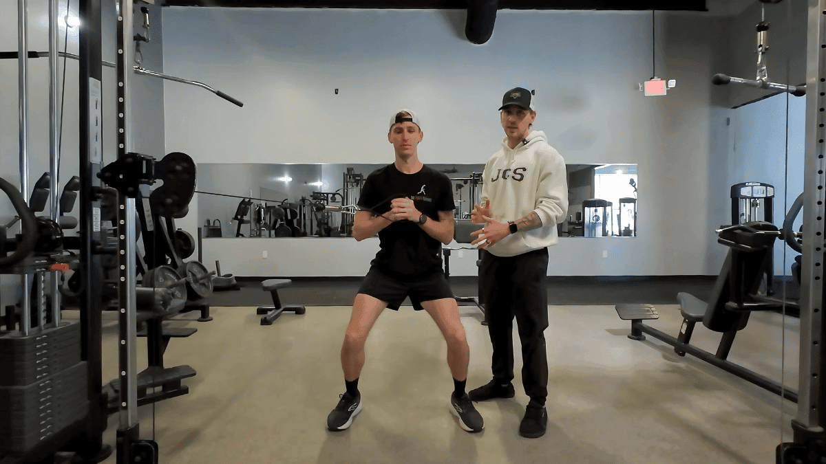 Two men in a gym demonstrating an exercise from the Runner Resistance Plan, with one in a squat position using a resistance band and the other providing instruction.