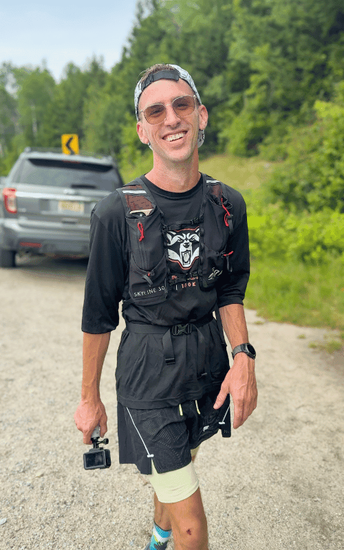 Smiling runner standing on a gravel roadside after completing the presidential traverse in one day, wearing a hydration vest and holding a small action camera, with a forested backdrop and parked SUV behind him.