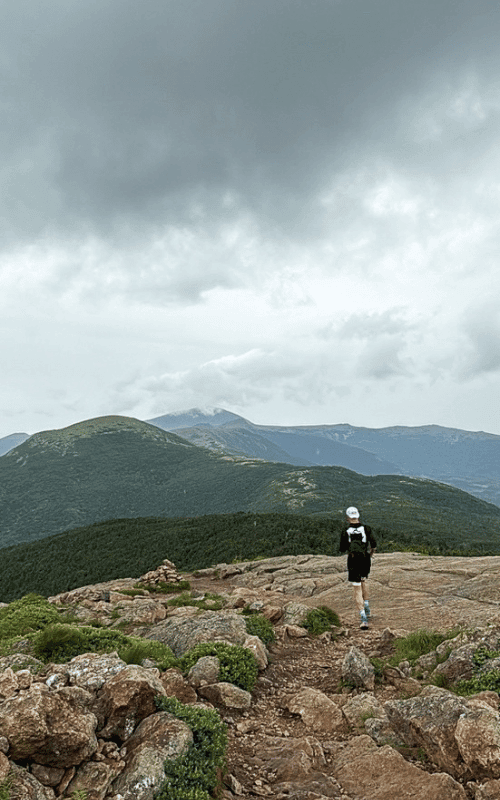 Solo runner running along a rocky alpine trail under moody gray skies, with Mount Washington and other peaks of the presidential traverse looming in the distance.