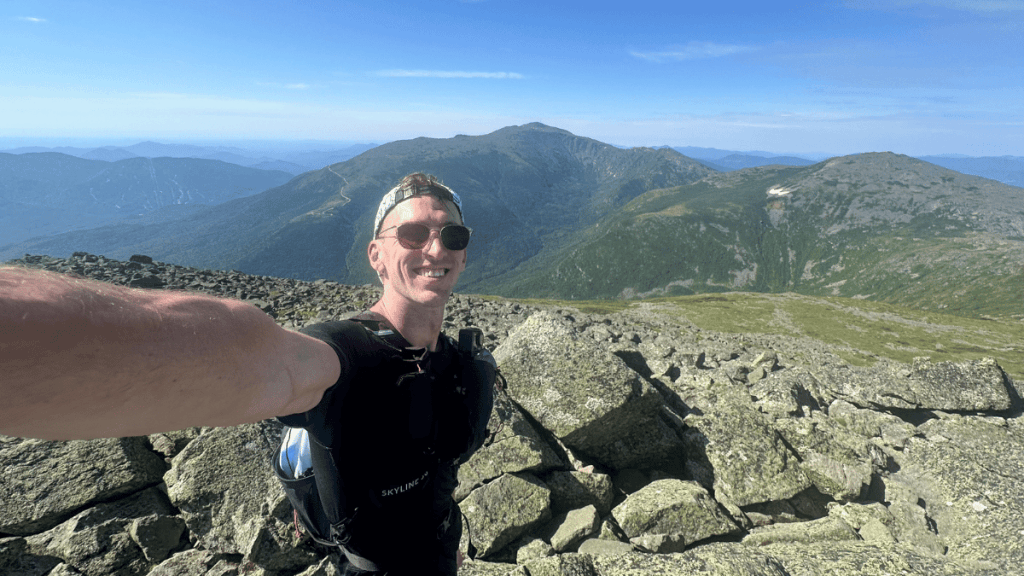 Runner pausing for a selfie on a boulder-strewn peak of Mount Adams, with expansive views of rugged terrain and Mount Washington in the background during a sunny day.