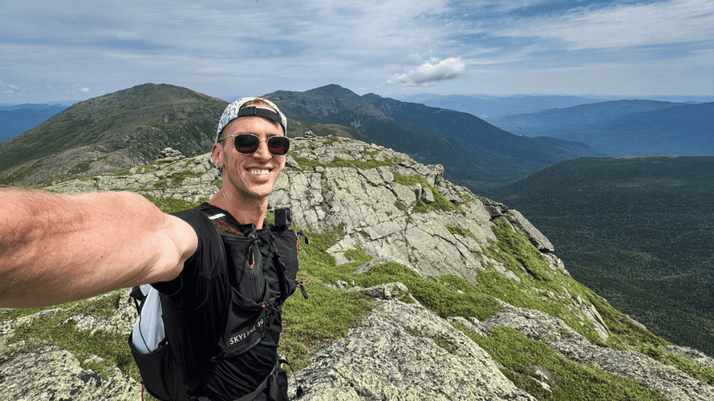 Runner smiling on a rocky outcrop on Mount Clay with rugged cliffs and several peaks behind him, capturing a scenic moment during the presidential traverse.