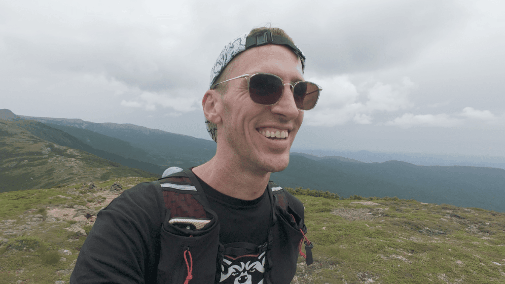 Close-up of a smiling hiker with windblown hair and sunglasses, hiking along a grassy alpine ridge on Mount Eisenhower with misty mountains fading into the background.