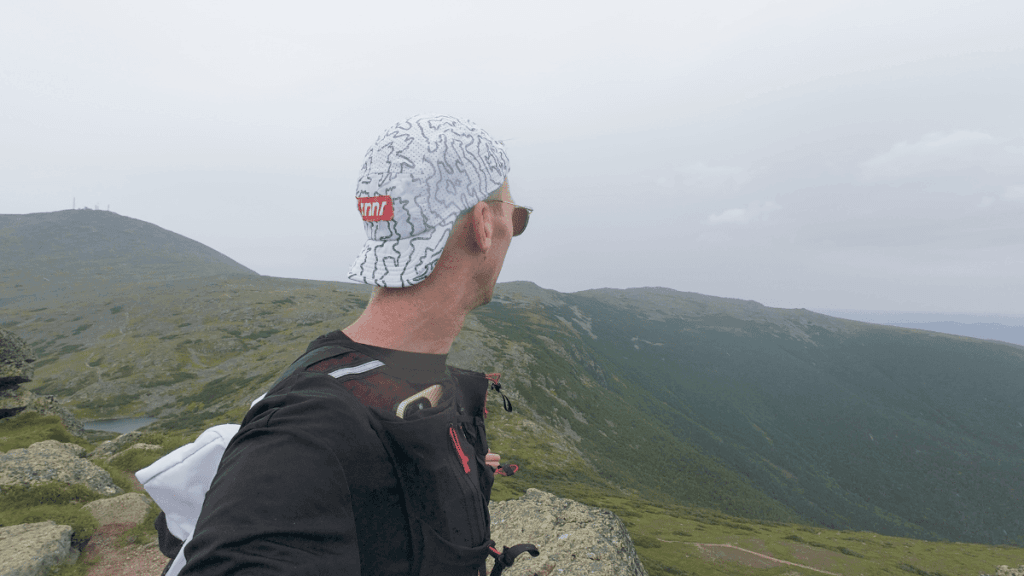 Runner wearing a patterned cap looking across a dramatic mountain valley from Mount Monroe, with overcast skies and expansive ridgelines in the distance.