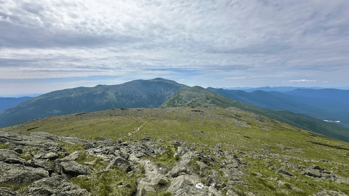 View from a rocky mountain ridge looking toward Mount Washington under a partly cloudy sky, with an alpine trail winding through the foreground on the Presidential Traverse.