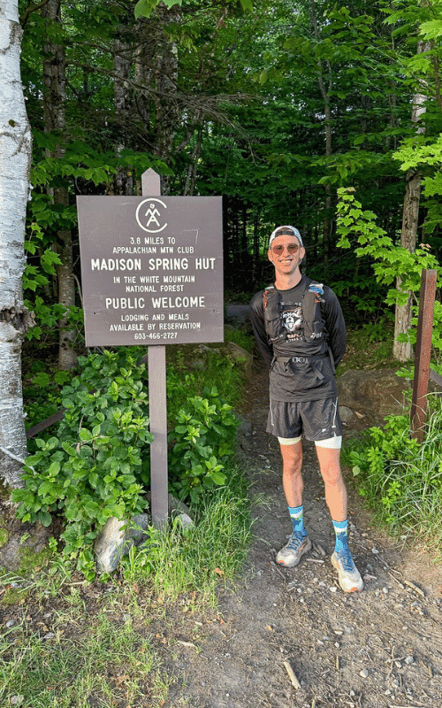 Runner standing next to a wooden trail sign in the White Mountain National Forest indicating 3.8 miles to Madison Spring Hut on the Appalachian Trail, surrounded by lush summer greenery.