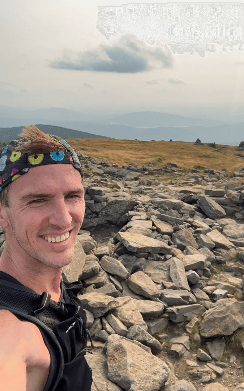 A smiling runner takes a selfie on the rocky summit of Mount Moosilauke, with sweeping mountain views and open alpine terrain in the background.