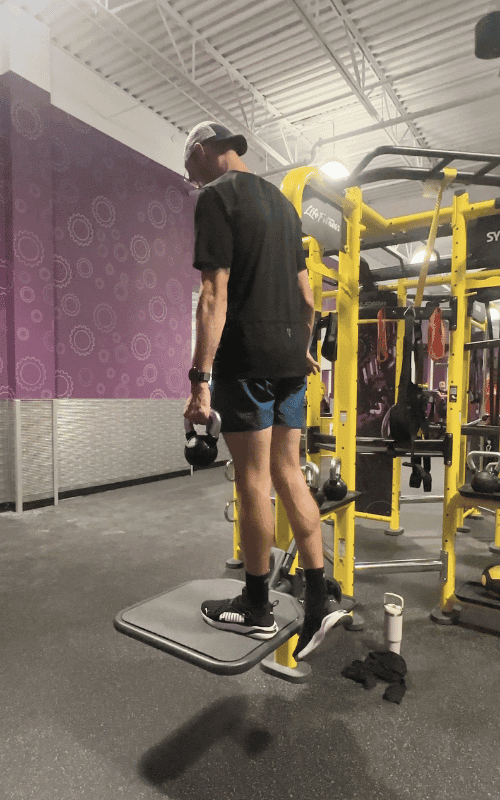 A runner standing on a raised platform performing a single-leg calf raise while holding a kettlebell, building lower leg strength important for strength training for runners.