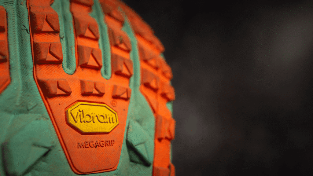 Close-up of the Vibram Megagrip outsole on the Altra Olympus 2.75, showing aggressive orange and teal lugs designed for superior traction. The yellow Vibram logo and "MEGAGRIP" text are clearly visible at the center of the sole.